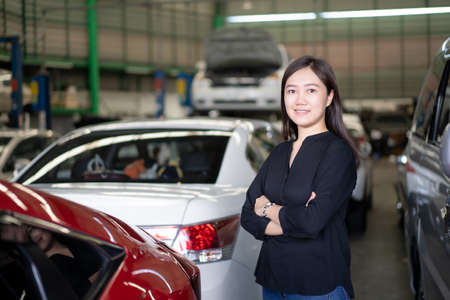 Portrait of beautiful Asian woman posing for photograph beside the car in garage, woman bring her vehicle to repair in garage. Satified Asian young woman standing beside the car in garage.の写真素材