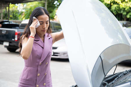 Unhappy beautiful Asian businesswoman standing in front of her broken vehicle beside the road, serious asian woman looking inside the broken car hood. Car accident and broken down.の写真素材