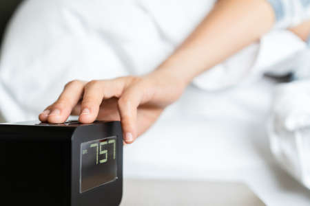 Lazy Asian young woman trying to snooze awaking digital alarm clock on the table beside the bed close up, woman wake up late in morning up to 8.00 am. Tried woman sleeping on the bed in bedroom.の写真素材