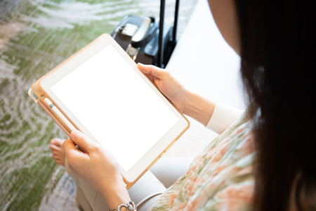 Mockup - blank screen tablet isolated with clipping. Happy Asian young woman tourist sitting on the bed in bedroom in hotel and using a digital tablet close up. Businesswoman tourist relaxing in hotelの写真素材