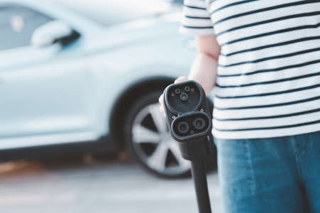 Happy Asian woman holding a DC - CCS type 2 EV charging connector at EV charging station, woman preparing an EV - electric vehicle charging connector for recharge a vehicle.の写真素材