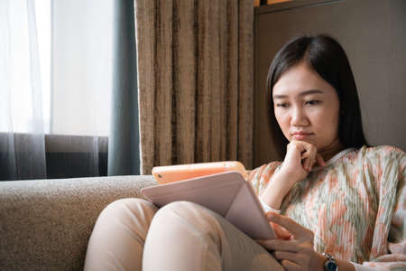 Asian young woman relaxing on the sofa bed in living room and using tablet for social network, woman using tablet for social media - video call to other people.の写真素材