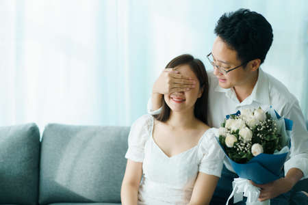 Happy Asian young couple in Valentine or anniversary stay together, man giving and surprise his wife a bouquet in their Valentine or Anniversary day. Husband and wife celebrating in Valentine day.の写真素材