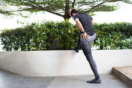 Happy Asian young sportsman doing a lower and upper body stretching before making an exercise in a park, stretching and workout concept. Sport man making a cardio and weight training workout.の写真素材