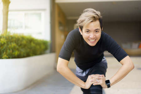 Happy Asian young sportsman doing a lower and upper body stretching before making an exercise in a park, stretching and workout concept. Sport man making a cardio and weight training workout.の写真素材