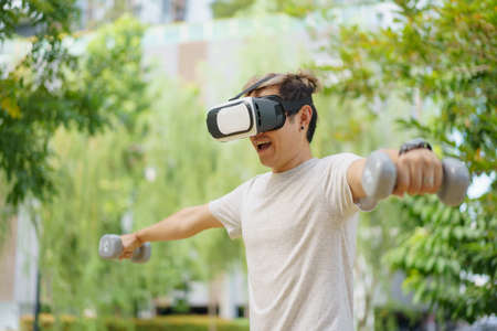 Active Asian sportsman doing a weight training exercise by using dumbbell and virtual reality - VR goggles platform for online training. The innovation of virtual reality or metaverse platform conceptの写真素材