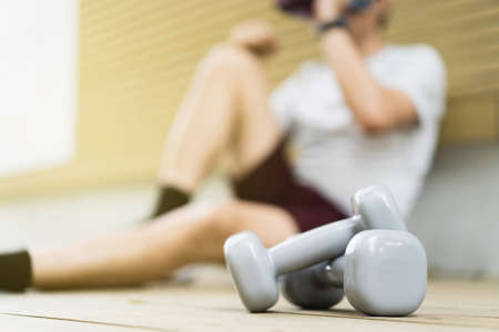 Asian young sportsman resting on the floor after weight training exercise, close up at a pair of dumbbell with blurred man in background with copyspace. Man taking a break after weight workout.の写真素材