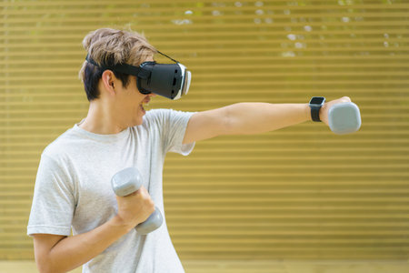 Active Asian sportsman doing a weight training exercise by using dumbbell and virtual reality - VR goggles platform for online training. The innovation of virtual reality or metaverse platform conceptの写真素材