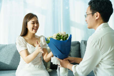 Happy Asian young couple in Valentine or anniversary stay together, man giving and surprise his wife a bouquet in their Valentine or Anniversary day. Husband and wife celebrating in Valentine day.の写真素材