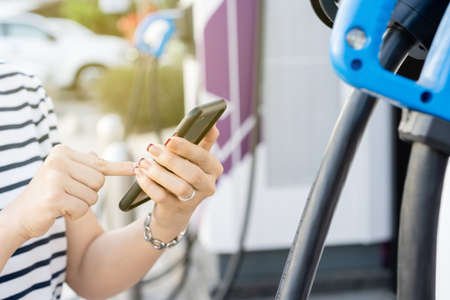 Happy Asian young woman using ev charging application on smartphone connect with the charging station, Bev or electric vehicle charging station. Woman refueling a battery at EV station.の写真素材