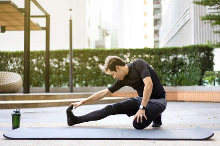 Happy Asian young sportsman doing a lower and upper body stretching before making an exercise in a park, stretching and workout concept. Sport man making a cardio and weight training workout.の写真素材