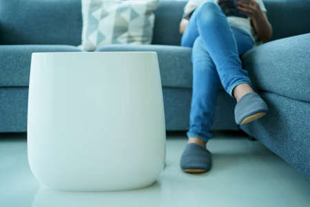 Happy Asian young woman turning on high efficiency air purifier while staying and reading a book in the living room. Woman using air purifier in her house.の写真素材