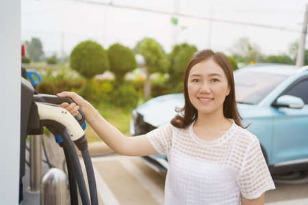 Happy Asian woman holding a DC - CCS type 2 EV charging connector at EV charging station, woman preparing an EV - electric vehicle charging connector for recharge a vehicle.の写真素材