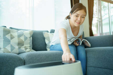 Happy Asian young woman turning on high efficiency air purifier while staying and reading a book in the living room. Woman using air purifier in her house.の写真素材