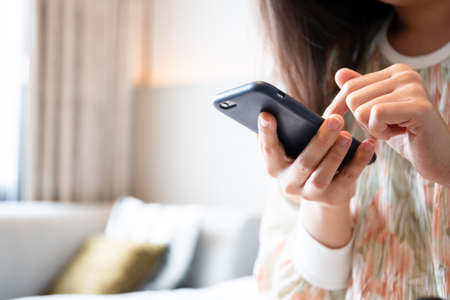 Asian young woman tourist sitting on the bed in bedroom in hotel and using smartphone close up. Happy Asian woman using smartphone for social media on smartphone while relaxing in the hotel.の写真素材