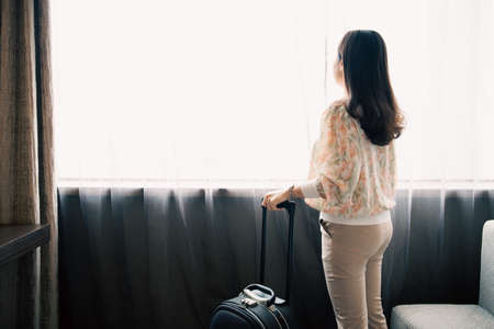 Rear and side view of Asian young woman smiling and opening window curtain in sky view hotel. Woman tourist open curtains and look outside the window after entered the room.の写真素材