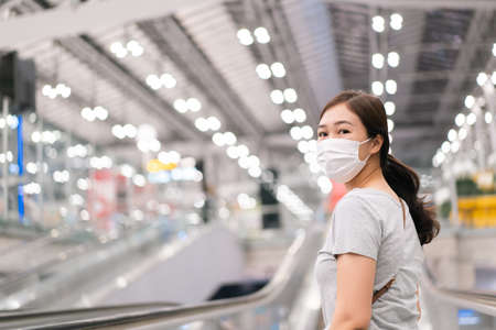 Beautiful Asian young woman with protective face mask looking and smiling to the camera while walking in the airport. Travel and tourist concept during the coronavirus or COVID-19 pandemic.の写真素材