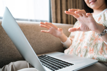 Happy Asian young woman making a video conference on laptop computer while living in the living room, cheerful Asian woman making a gesture or hands sign in video call.の写真素材