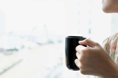 Young Asian woman holding a coffee cup near the window, woman drinking a coffee. Happy woman drinking hot coffee in a mug in the morning close up with copyspace.の写真素材