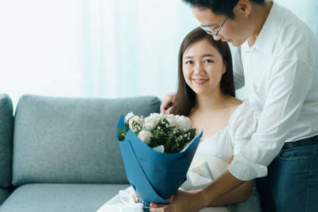 Happy Asian young couple in Valentine or anniversary stay together, man giving and surprise his wife a bouquet in their Valentine or Anniversary day. Husband and wife celebrating in Valentine day.の写真素材