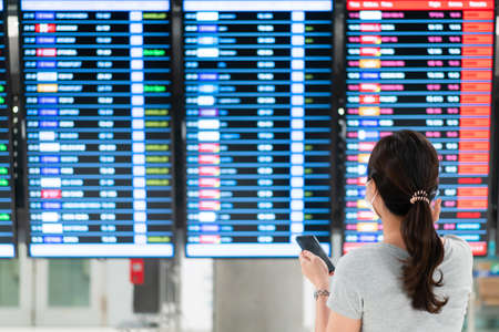 Beautiful Asian young woman with protective face mask standing in the airport and looking at the flight information. Woman wearing a face mask while traveling the coronavirus - COVID-19 pandemic.の写真素材