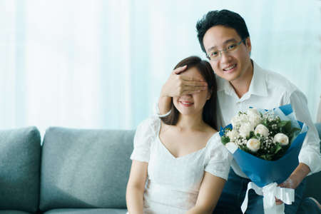 Happy Asian young couple in Valentine or anniversary stay together, man giving and surprise his wife a bouquet in their Valentine or Anniversary day. Husband and wife celebrating in Valentine day.の写真素材