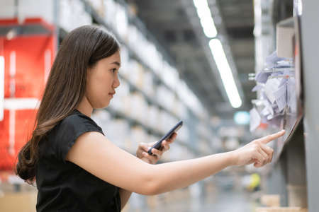 Asian young woman wearing a protective face mask searching on a smartphone and looking for a goods and products on a shelf, self-service concept. Cheerful woman walking in a warehouse.の写真素材