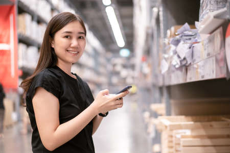 Asian young woman wearing a protective face mask searching on a smartphone and looking for a goods and products on a shelf, self-service concept. Cheerful woman walking in a warehouse.の写真素材