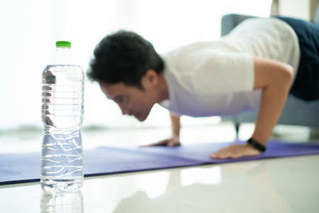 Unrecognizable Asian young man making a push up workout at home with a bottle of water on the floor,...
