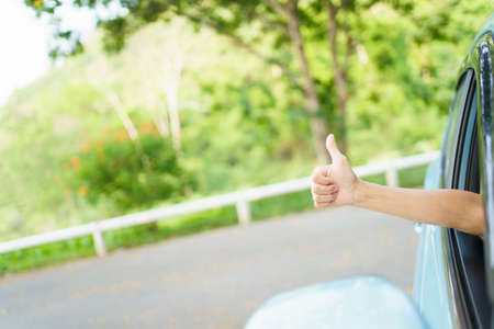 Unrecognizable Asian woman driver showing a thumb up outside the vehicle while driving a car. Happy woman driver or passenger showing her thumb up during in the car. Safety trip and freedom concept.の写真素材