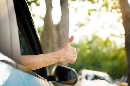 Unrecognizable Asian woman driver showing a thumb up outside the vehicle while driving a car. Happy woman driver or passenger showing her thumb up during in the car. Safety trip and freedom concept.の写真素材