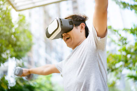 Active Asian sportsman doing a weight training exercise by using dumbbell and virtual reality - VR goggles platform for online training. The innovation of virtual reality or metaverse platform conceptの写真素材