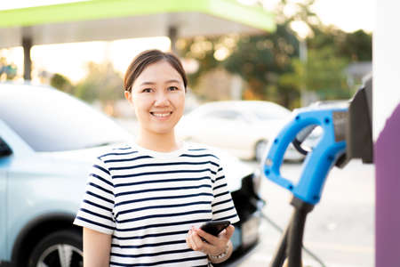 Happy Asian young woman using ev charging application on smartphone connect with the charging station, Bev or electric vehicle charging station. Woman refueling a battery at EV station.の写真素材