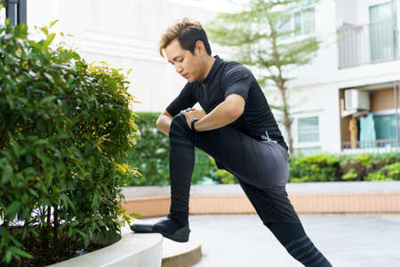 Happy Asian young sportsman doing a lower and upper body stretching before making an exercise in a park, stretching and workout concept. Sport man making a cardio and weight training workout.の写真素材