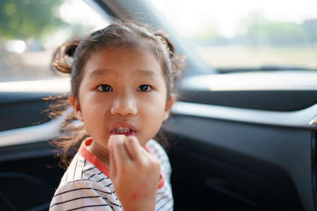 Little Asian young girl staying in the car and eating fried potato chips, lovely girl enjoy eating snack while traveling by the car.の写真素材