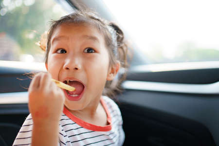 Little Asian young girl staying in the car and eating fried potato chips, lovely girl enjoy eating snack while traveling by the car.の写真素材