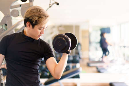 Active Asian sportsman doing a weight exercise in a gym - fitness by lifting up a dumbbell, man sitting on a exercise bench and lift up dumbbells. Sportsman making a shoulder and arms exercise.の写真素材
