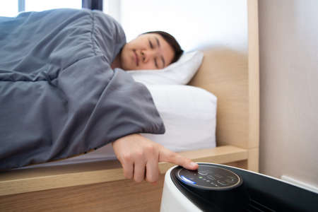 Asian sleepy woman trying to press a button on the portable air purifier machine in a bedroom. Lazy woman sleeping on the bed and trying to turn the air purifier on. Wellbeing and healthy lifestyle.の写真素材