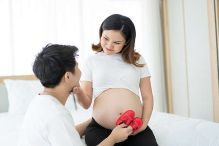 Asian ethnicity happy pregnant woman showing a baby red shoes on her big belly close up with copyspace. Touching emotion of love of mother and father. Being maternity and parenthood concept.の写真素材