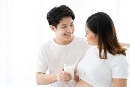 Lovely Asian couple taking care together in their house, husband taking care his pregnant wife by giving a fresh milk. Wellness and health care in pregnancy woman and unborn child in tummy.の写真素材
