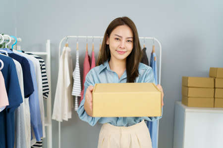 Happy Asian young woman showing a cardboard parcel box to camera, online selling or ecommerce concept. Female online seller sells a clothes via online marketplace platform.の写真素材