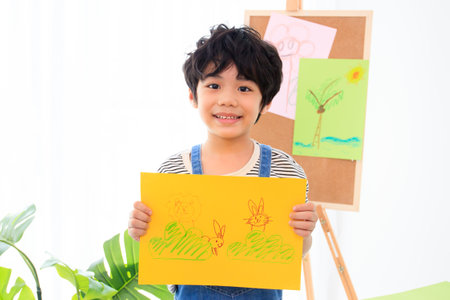 Happy little Asian boy showing his painting - drawing art on a paper to camera. Education and development in the elementary or preschool age concept. Kindergarten children portrait.の写真素材