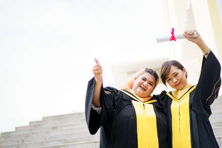 Happy Asian young women in the master degree gown showing a diploma in their hand close up. Portrait of confident college students in gown in the graduation ceremony.の写真素材