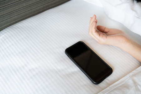 Lazy Asian young woman trying to snooze awaking alarm clock in smartphone close up, woman wake up late in morning up to 8.00 am. Tried woman sleeping on the bed in bedroom.の写真素材