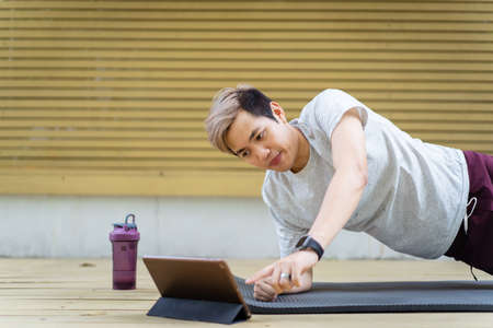 Active sportsman doing a plank exercise and making a video call with a friend and online sport training coach online. Healthy sport man making a body building workout with online instructor.の写真素材