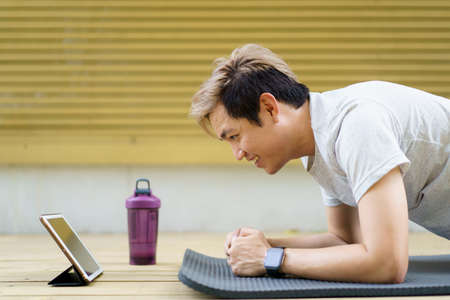 Active sportsman doing a plank exercise and making a video call with a friend and online sport training coach online. Healthy sport man making a body building workout with online instructor.の写真素材