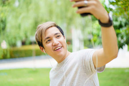 Handsome Asian young sportsman posing and smiling to the camera while drinking water, man listening a music on smartphone while practicing running workout. Happy Asian athlete smiling.の写真素材