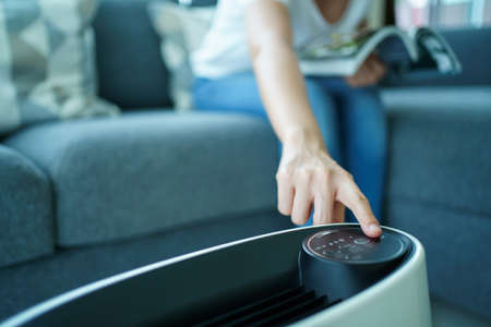 Happy Asian young woman turning on high efficiency air purifier while staying and reading a book in the living room. Woman using air purifier in her house.の写真素材