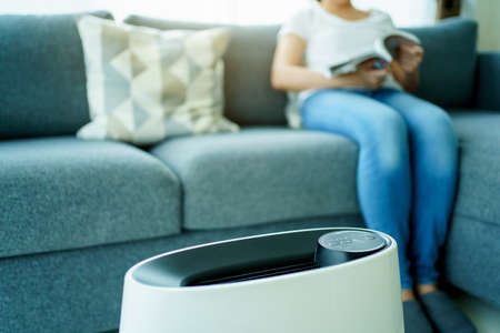 Happy Asian young woman turning on high efficiency air purifier while staying and reading a book in the living room. Woman using air purifier in her house.の写真素材