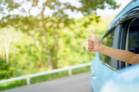 Unrecognizable Asian woman driver showing a thumb up outside the vehicle while driving a car. Happy woman driver or passenger showing her thumb up during in the car. Safety trip and freedom concept.の写真素材